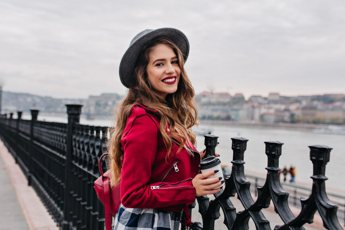 Pretty curly woman with bright makeup enjoying city view from bridge in autumn day. Lovely brunette woman in red jacket spending time on embankment during rest in Europe.