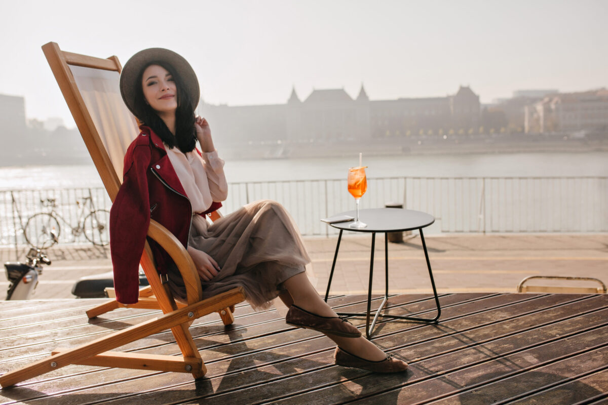 Smiling brunette girl sitting on recliner enjoying vacation in Europe. Portrait of well-dressed lady in good mood chilling in outdoor cafe with cocktail.