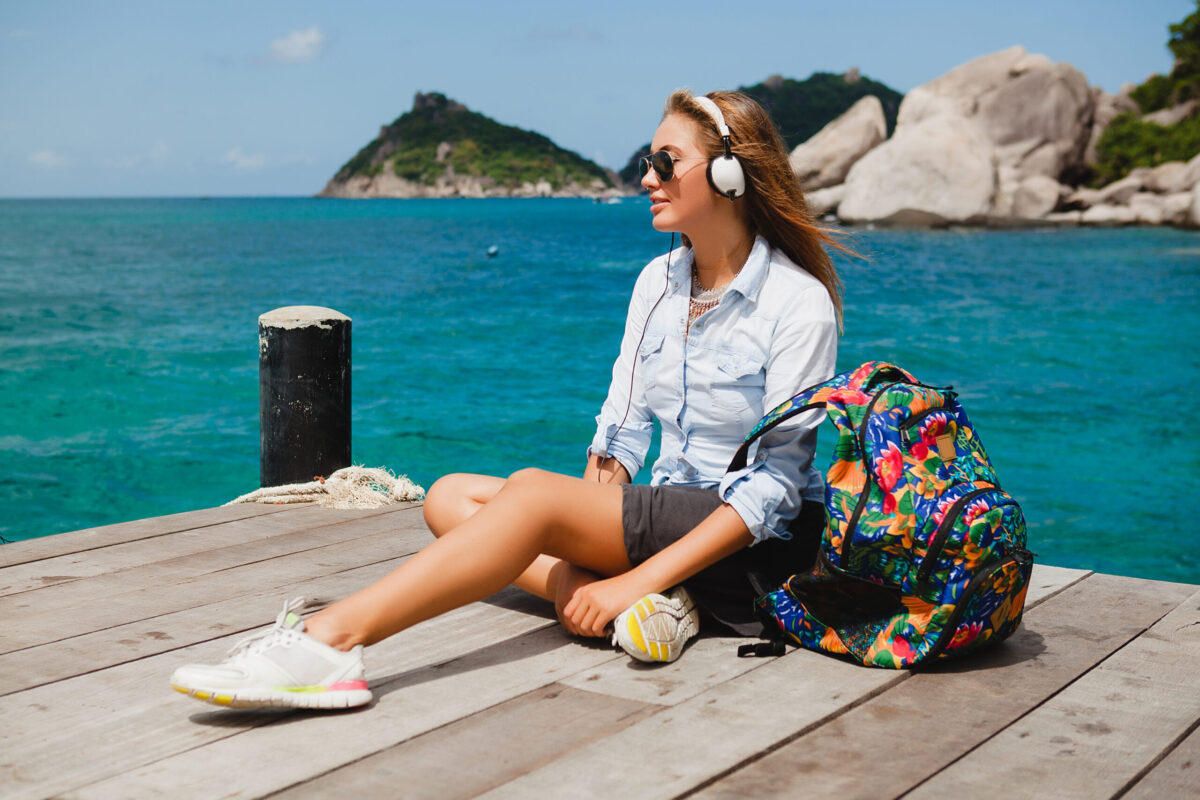 young stylish hipster woman traveling around the world, sitting on the pier, aviator sunglasses, headphones, listening to music, vacation, backpack, denim shirt, happy, tropical island lagoon